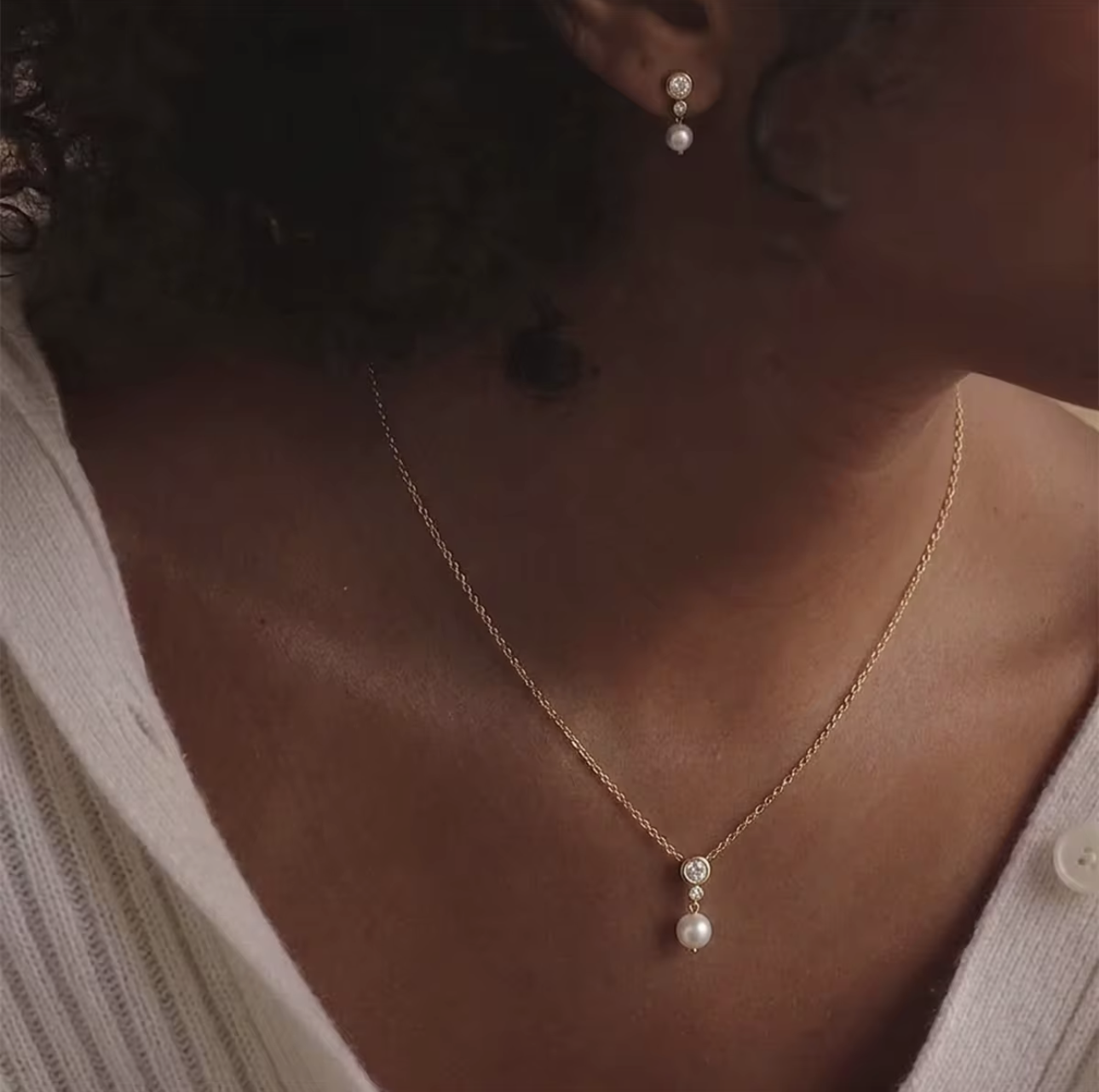 A close-up lifestyle shot of a model wearing a delicate gold chain necklace with a double pearl pendant, paired with a matching pearl stud earring and a white ribbed top.