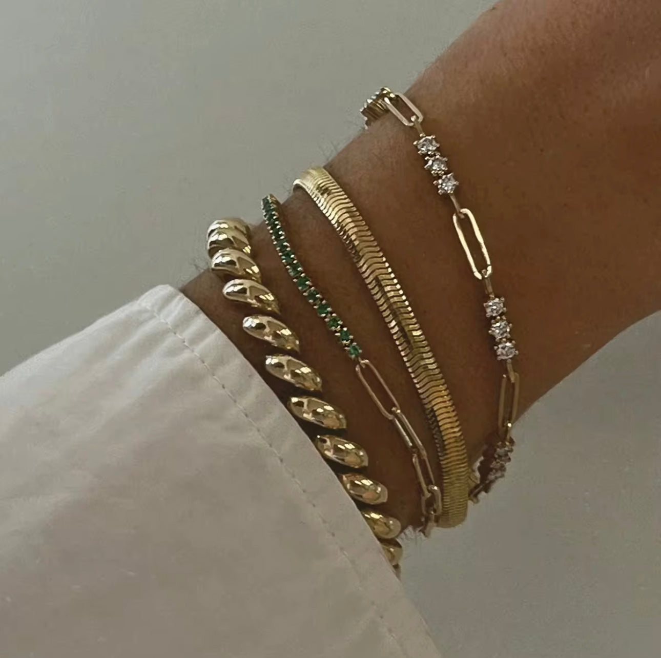 multiple gold bracelets being worn on a wirst with a white sleeve and neutral background
