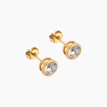 A pair of gold-colored stud earrings with  a round clear gemstone, pictured on a white background