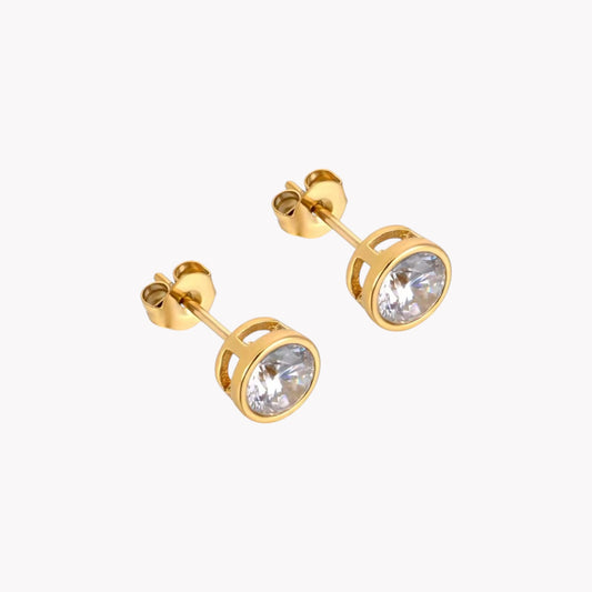 A pair of gold-colored stud earrings with  a round clear gemstone, pictured on a white background
