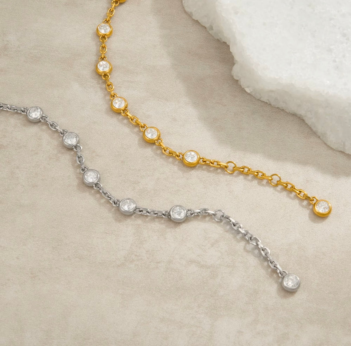 Two delicate chain necklaces with small round clear stones, one in yellow gold and one in silver, arranged on a light beige textured surface with a white stone prop in the corner.