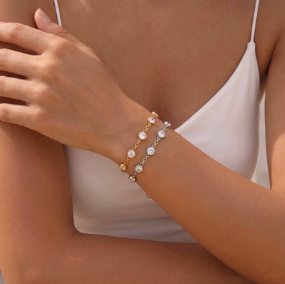 A close-up of a model's wrist wearing two delicate bracelets: one gold chain with round clear stones, and one silver chain with oval clear stones, styled with a white camisole.