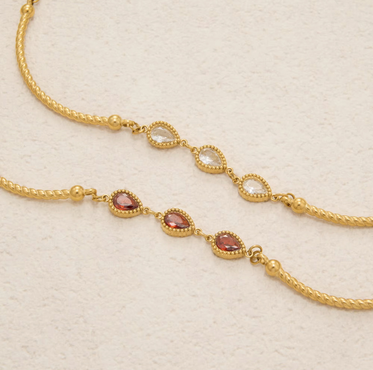 Gold-toned chain jewelry with three rows of teardrop-shaped clear stones and three rows of oval reddish-pink stones, displayed on a textured off-white background.