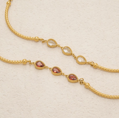 Gold-toned chain jewelry with three rows of teardrop-shaped clear stones and three rows of oval reddish-pink stones, displayed on a textured off-white background.
