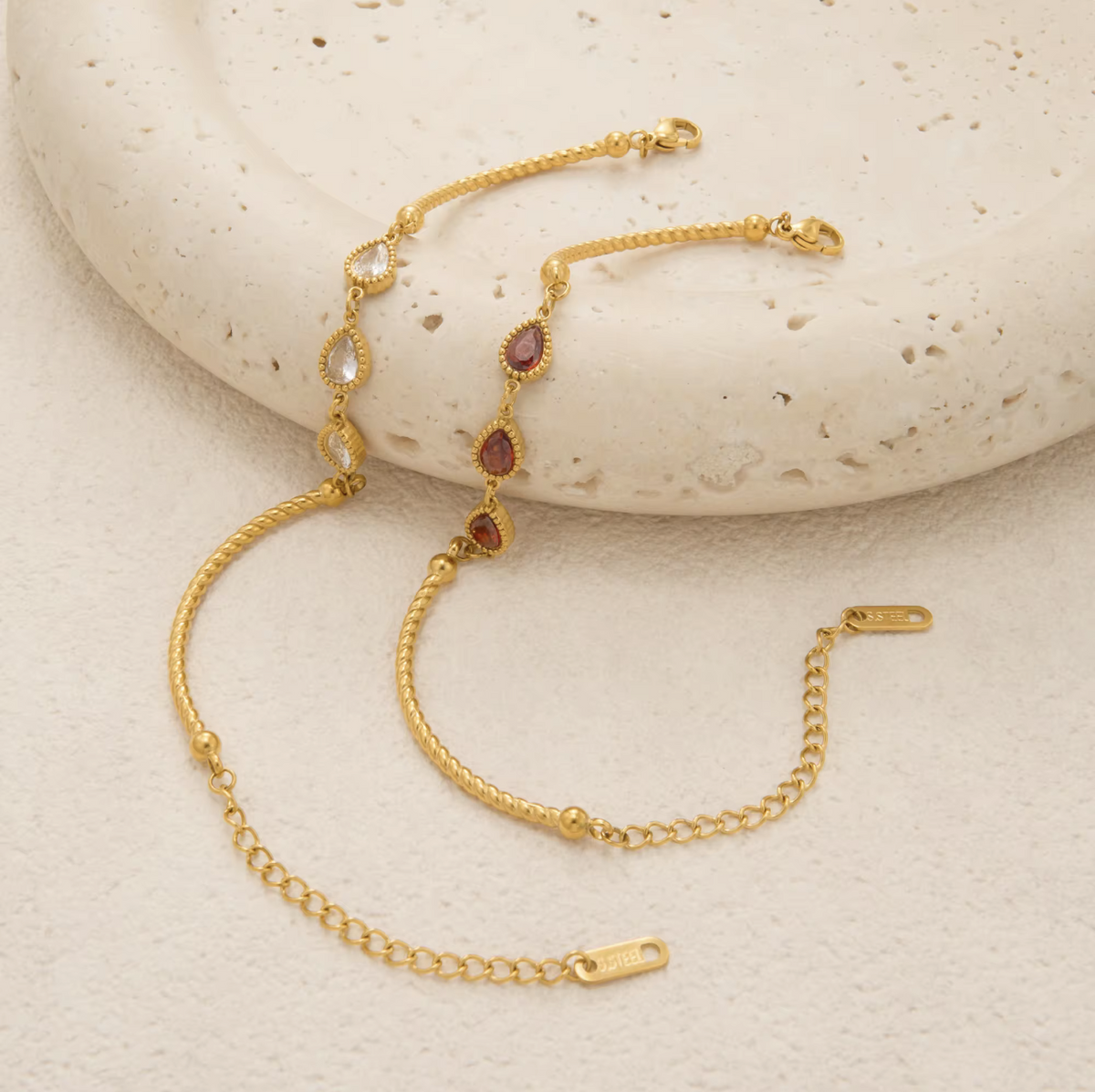 Two delicate gold chain anklets with decorative oval and marquise-cut stones, one clear and one reddish-brown, displayed on a textured beige stone slab.