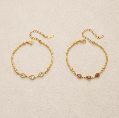 Two gold-toned delicate bracelets displayed on a textured off-white background. The left bracelet features a twisted rope design with a central infinity loop set with small clear stones.