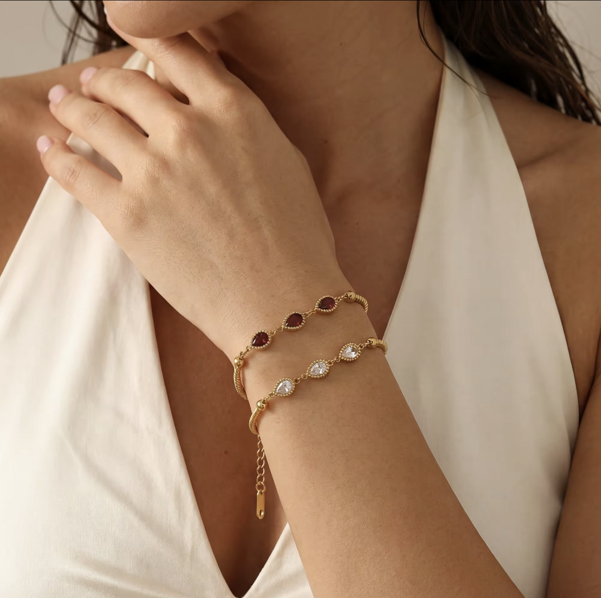 A close-up of a woman's wrist wearing two delicate gold bracelets. One bracelet features small oval dark red gemstones, and the other features clear marquise-cut stones, both on a gold-toned band with an extender chain.