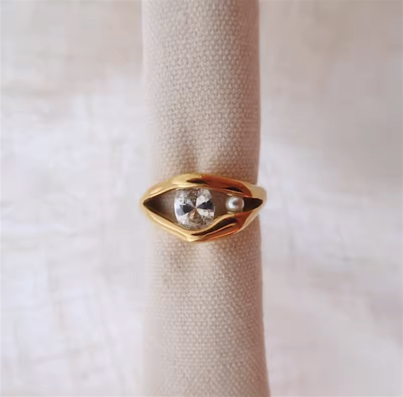 Gold ring with a diamond and pearl on a beige cushion against a light background