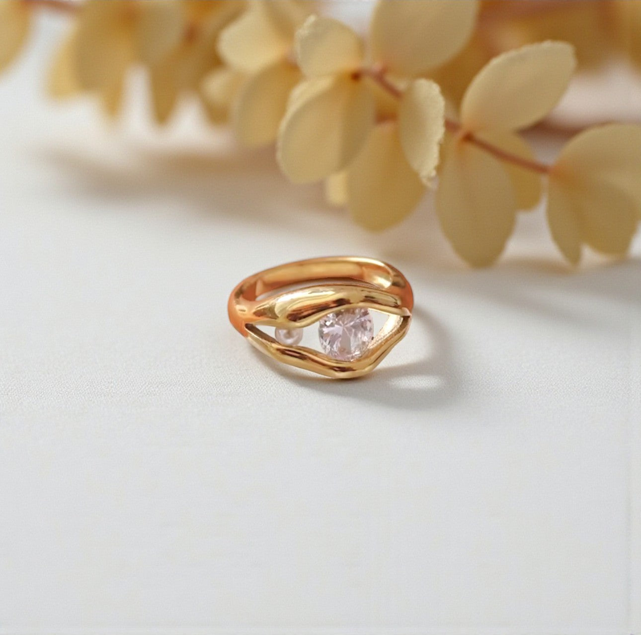 Gold ring with a clear gemstone and pearl on a light background with yellow leaves.
