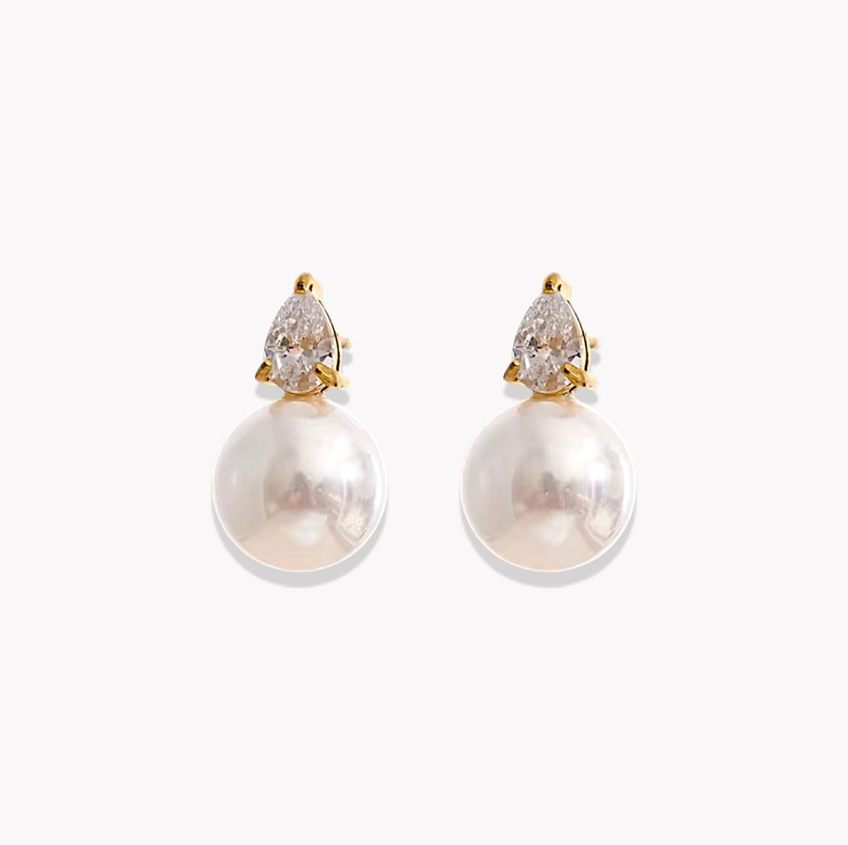 A pair of gold drop earrings featuring a pear-cut clear gemstone in a prong setting above a large, lustrous white pearl, shown on a light gray background.
