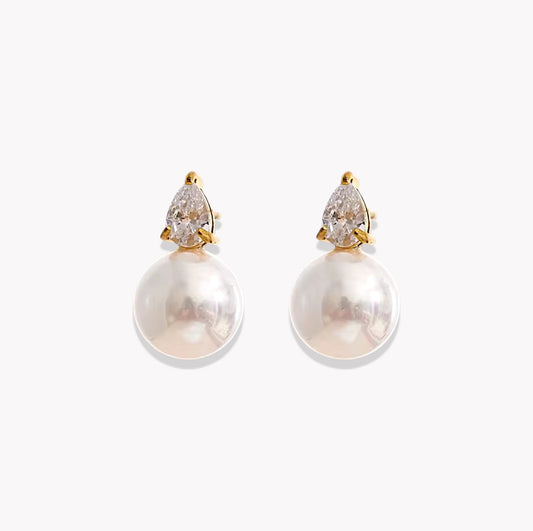 A pair of gold drop earrings featuring a pear-cut clear gemstone in a prong setting above a large, lustrous white pearl, shown on a light gray background.