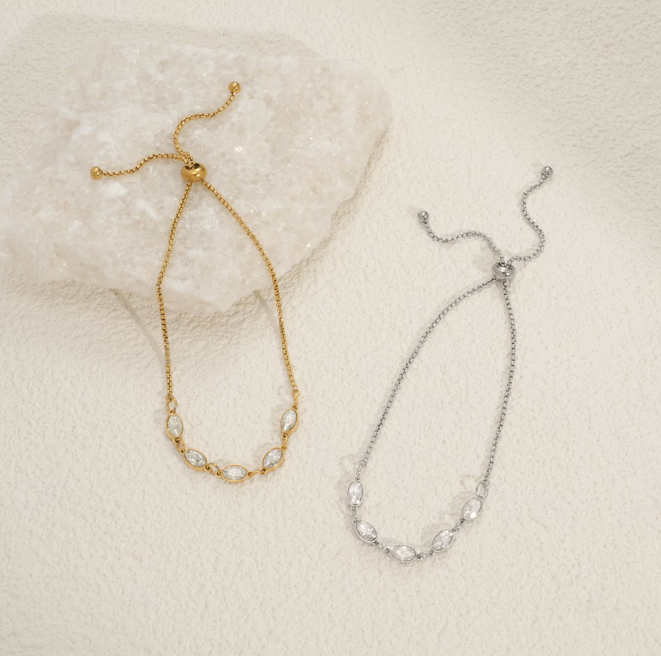 Gold and silver bracelets each featuring five oval shaped clear gemstones on a textured beige surface