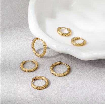 Five pairs of textured gold hoop earrings arranged on a light gray surface beside a white ceramic dish.