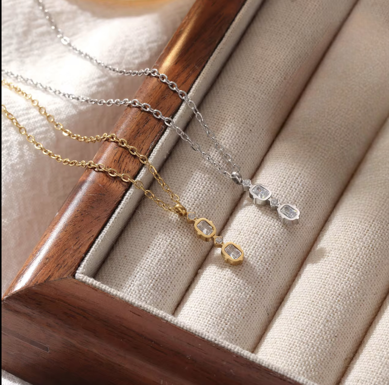 Two necklaces, one in gold and one in silver on a wooden stand with a beige fabric background.