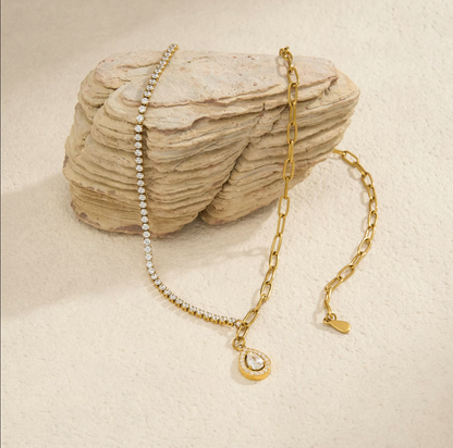 A gold chain necklace with a small teardrop-shaped pendant set with a clear stone, draped over a textured beige rock on a light beige background.