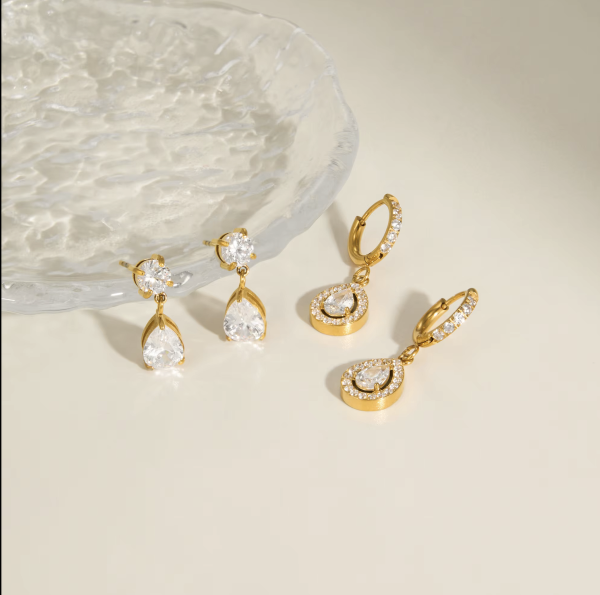 Four gold-tone drop earrings with clear gemstones, arranged on a light beige surface next to a textured clear glass dish.