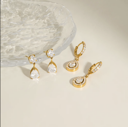 Four gold-tone drop earrings with clear gemstones, arranged on a light beige surface next to a textured clear glass dish.