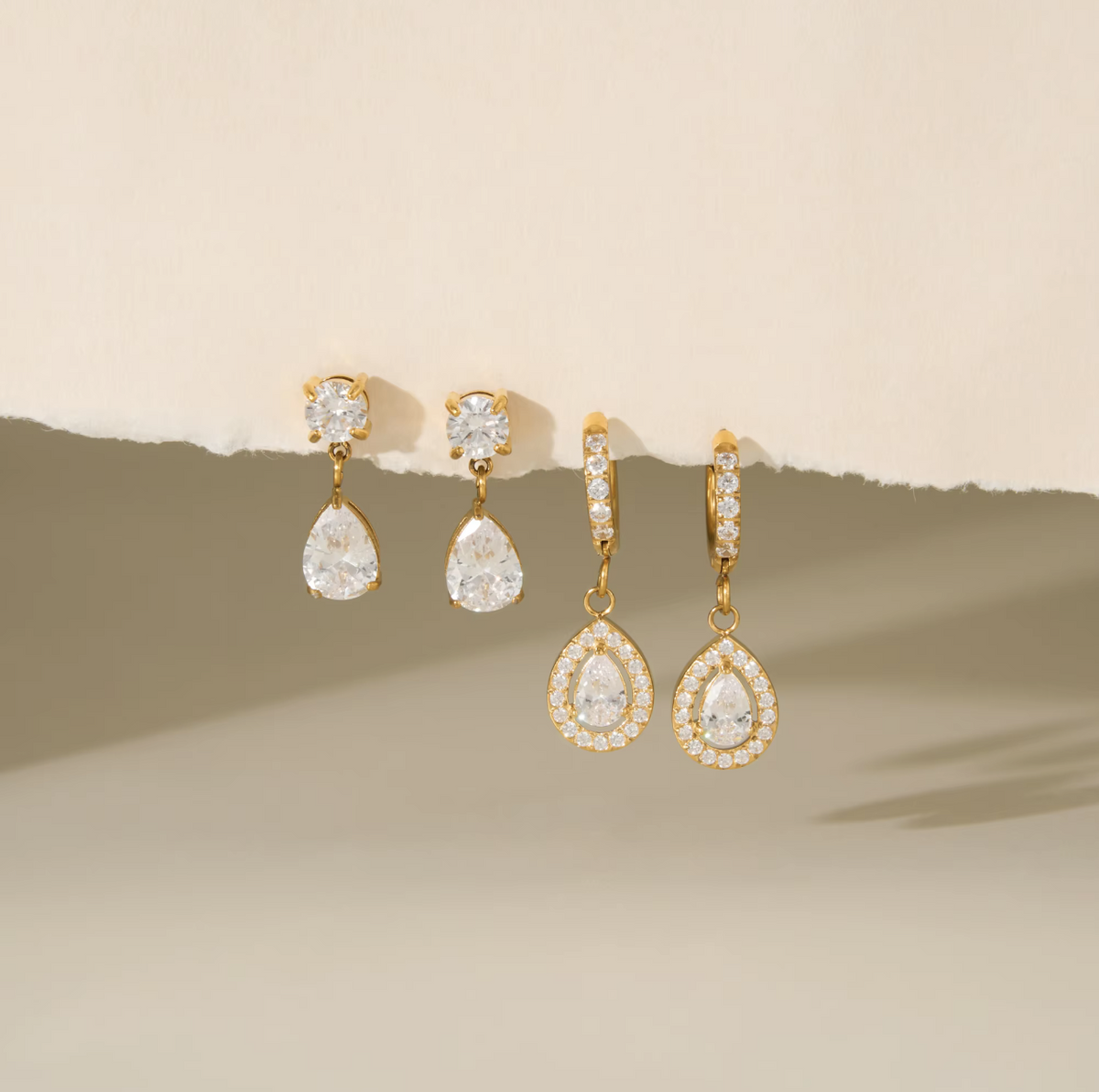 Four gold-tone drop earrings with clear teardrop-shaped gemstones and small clear stones in halo settings, arranged against a warm beige background with a torn-paper effect.