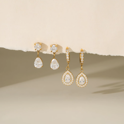 Four gold-tone drop earrings with clear teardrop-shaped gemstones and small clear stones in halo settings, arranged against a warm beige background with a torn-paper effect.
