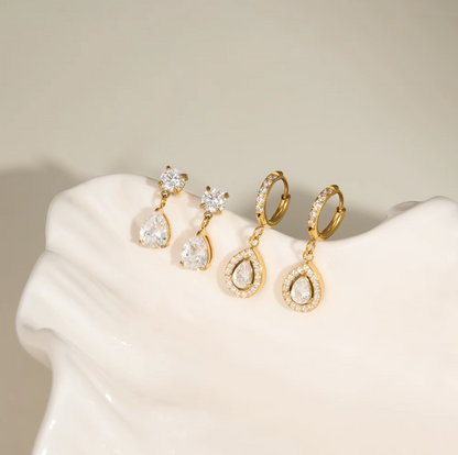 Three pairs of gold and crystal drop earrings displayed on a white ceramic jewelry stand against a warm beige background.