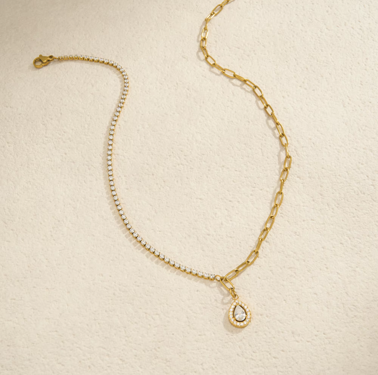 A gold chain anklet featuring a delicate pavé chain section, a larger curb chain section, and a teardrop-shaped pendant with a clear crystal surrounded by a gold border, displayed on a textured cream background.