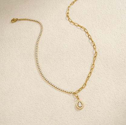 A gold chain anklet featuring a delicate pavé chain section, a larger curb chain section, and a teardrop-shaped pendant with a clear crystal surrounded by a gold border, displayed on a textured cream background.