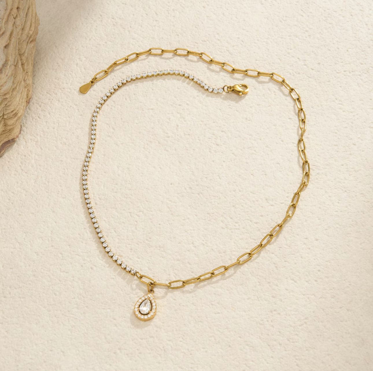 A delicate gold chain necklace with a small teardrop-shaped clear crystal pendant, displayed on a textured cream surface.
