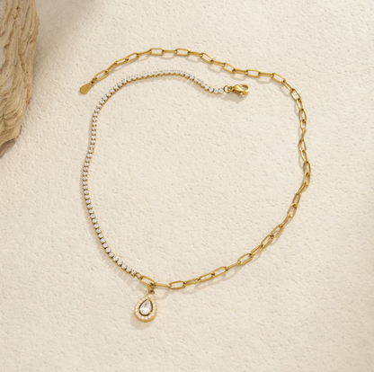 A delicate gold chain necklace with a small teardrop-shaped clear crystal pendant, displayed on a textured cream surface.