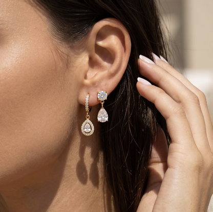 A close-up of a woman's ear wearing a pair of gold drop earrings with clear teardrop-shaped gemstones and small clear stones, with her hand near her face.
