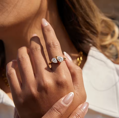 Close-up of a hand wearing a gold ring with two diamonds, held against a blurred background.