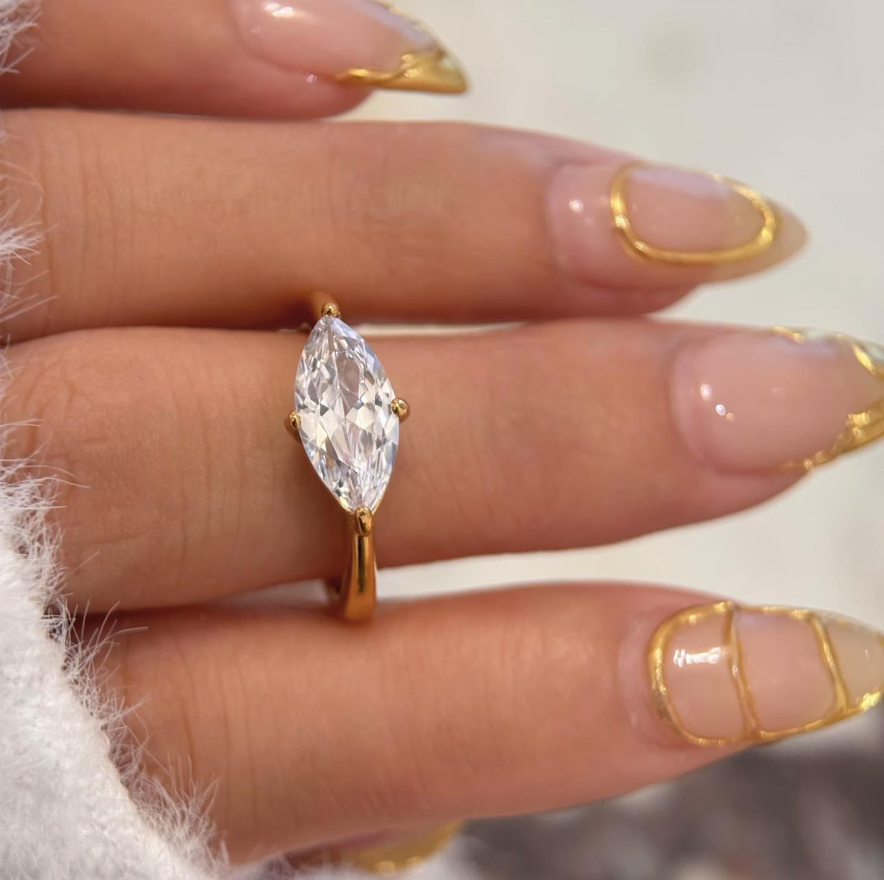 Gold ring featuring a large marquise shaped clear gemstone on a finger with manicured nails