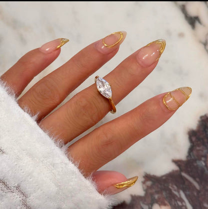 Gold ring featuring a large marquise shaped clear gemstone shown on a finger with manicured nails and marble background
