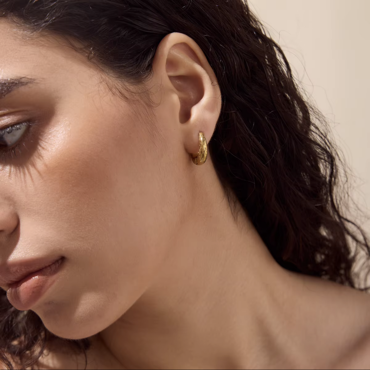 A close-up of a woman's ear wearing a small textured gold hoop earring against a warm beige background.