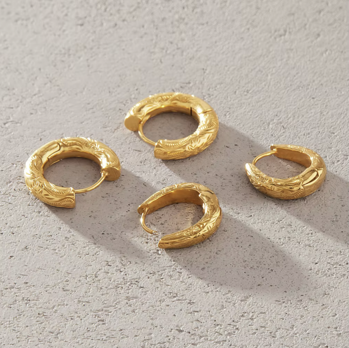 Four textured gold huggie hoop earrings arranged on a light gray concrete surface.