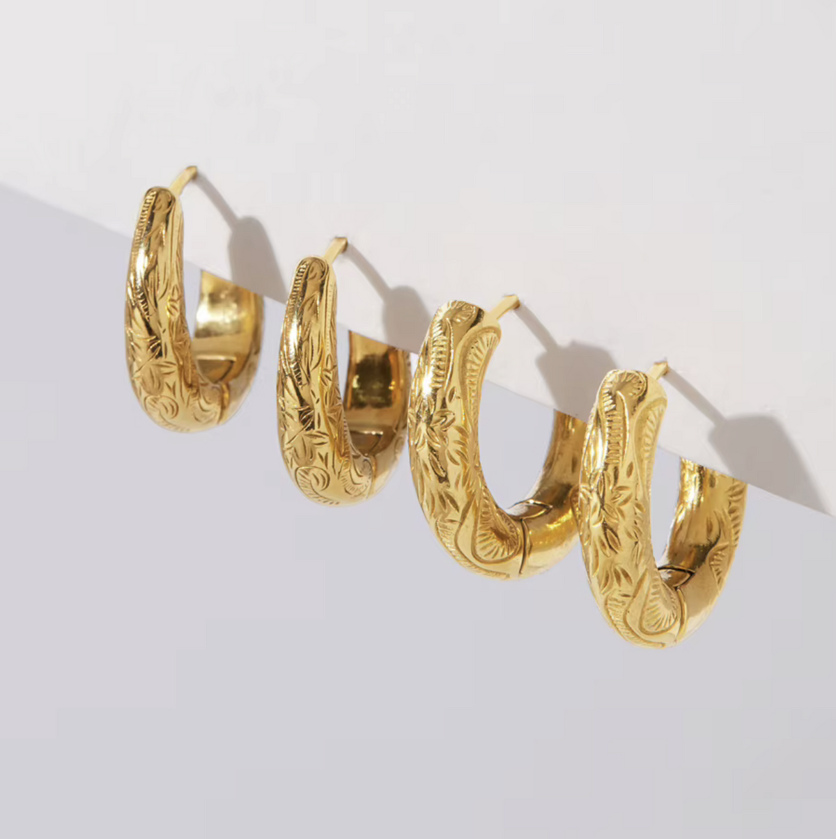 Four gold huggie hoop earrings with intricate floral engraving, arranged diagonally on a white surface.