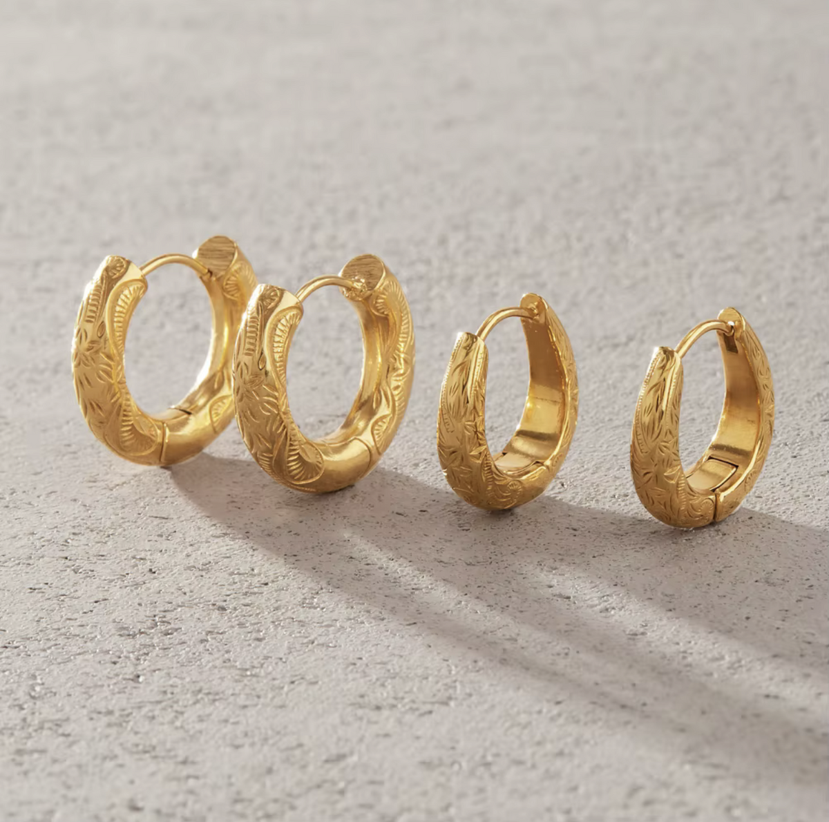 Four gold huggie hoop earrings with textured leaf engravings, arranged in descending size order on a light gray textured surface.