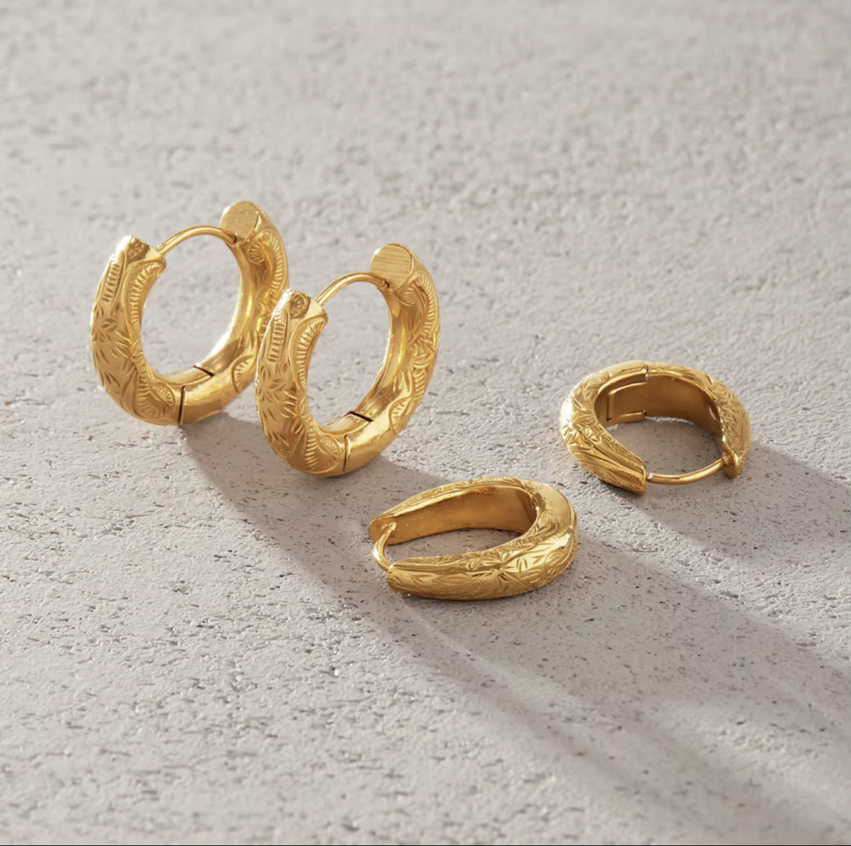 Three textured gold huggie hoop earrings arranged on a light gray concrete surface with soft shadows.