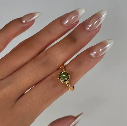 Gold ring with a green gemstone on a hand with long, manicured nails against a neutral background