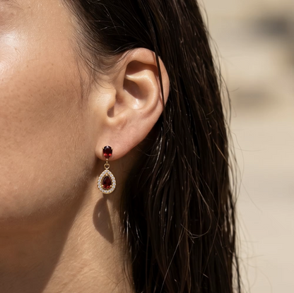 A close-up of a woman's ear wearing a gold drop earring with a dark red top bead and a teardrop-shaped red stone surrounded by small clear stones.