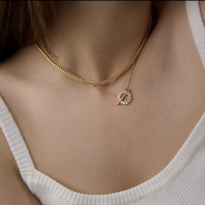 A close-up of a model's neck and chest wearing a double-layered gold necklace. The top layer is a thicker curb chain, and the bottom layer is a delicate chain with a small crescent moon pendant encrusted with clear rhinestones.