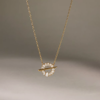 A delicate gold chain necklace featuring a circular pendant adorned with clear, faceted gemstones and crossed by a thin gold bar, set against a soft, neutral gray background.