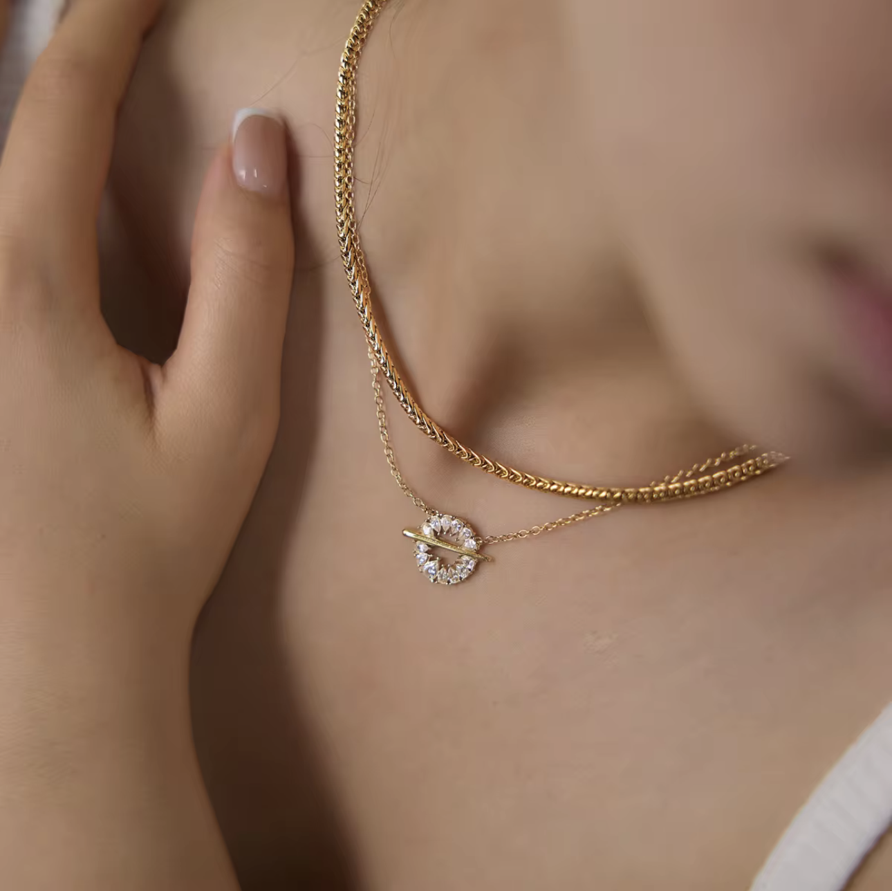 A close-up lifestyle shot of a model wearing a delicate gold necklace with a circular crystal-encrusted pendant and a second layered chain.