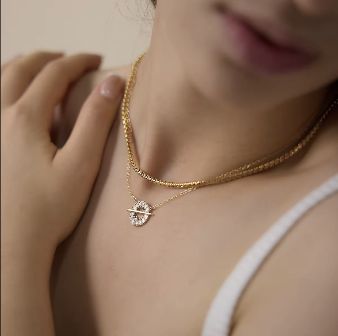 A close-up lifestyle shot of a model wearing a delicate double-layered necklace. The necklace features a gold chain with a small gold bar and a central cluster of clear, sparkling stones.