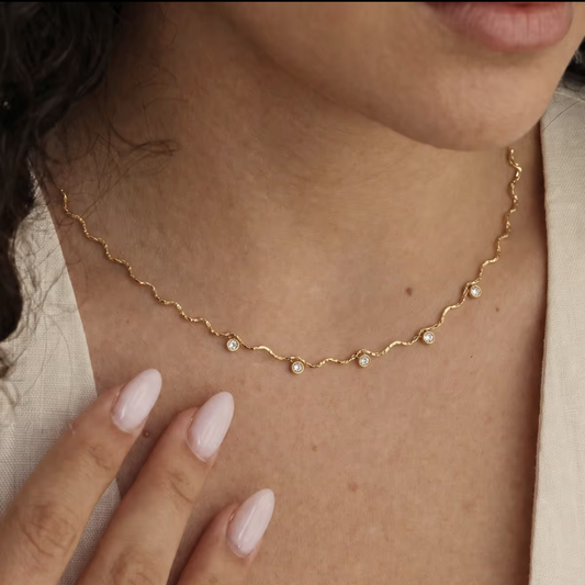 A close-up of a woman's neck and chest wearing a delicate gold chain necklace with small white pearl-like beads spaced along the chain, paired with a light beige top.