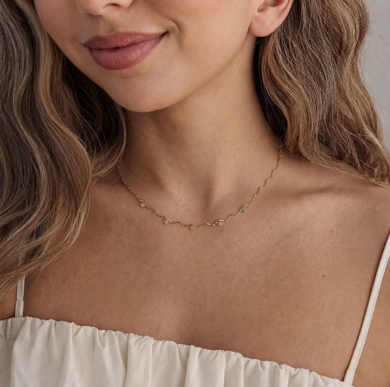 A close-up lifestyle shot of a model wearing a delicate gold chain necklace with small, clear, faceted gemstones spaced along the chain. The model is wearing a cream-colored off-the-shoulder top, and her hair is styled in soft waves.