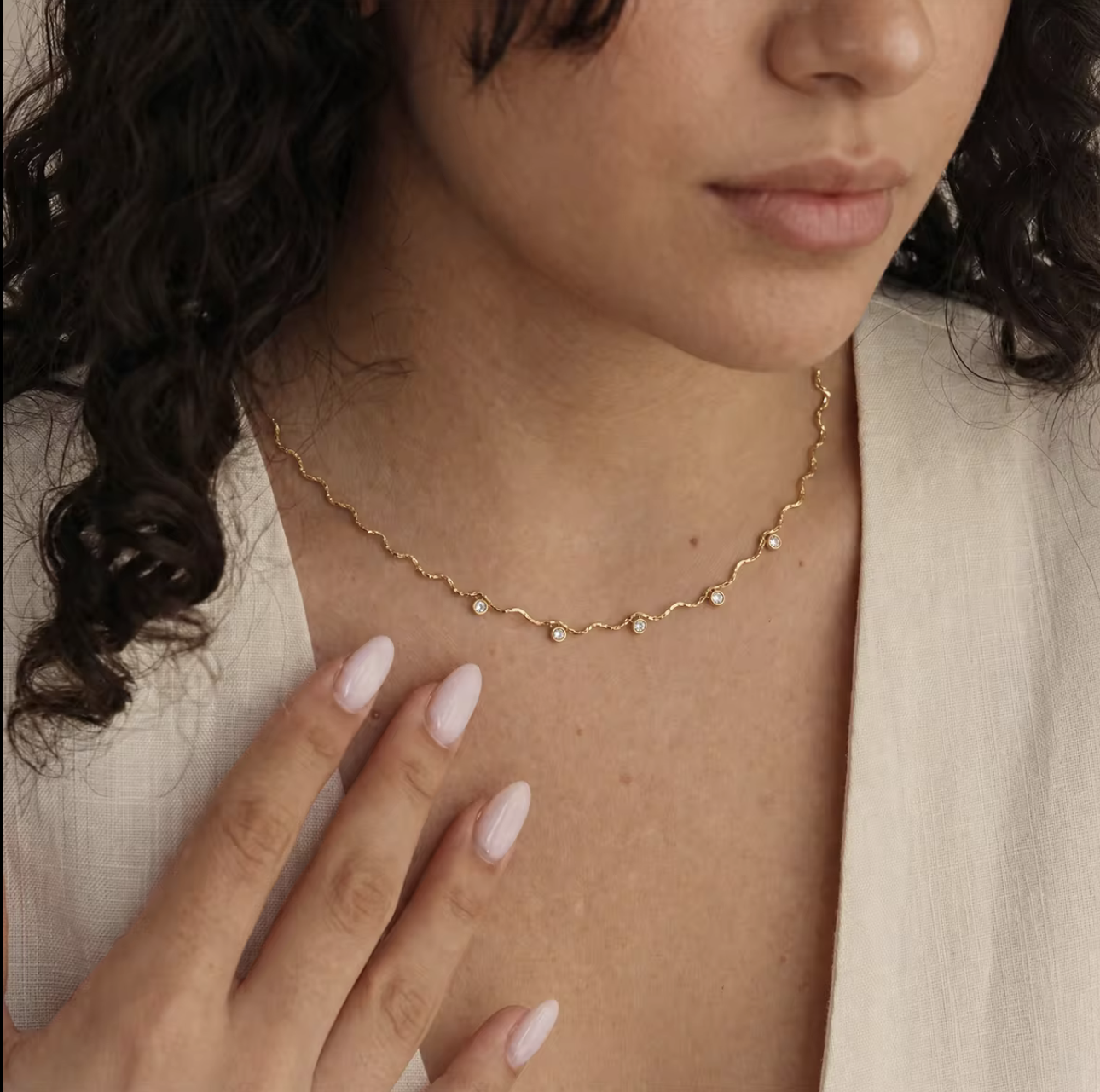 A close-up of a woman wearing a delicate gold chain necklace with small white pearl-like beads, styled with a cream-colored top.