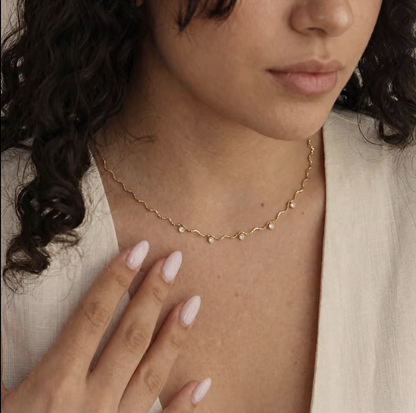 A close-up of a woman wearing a delicate gold chain necklace with small white pearl-like beads, styled with a cream-colored top.