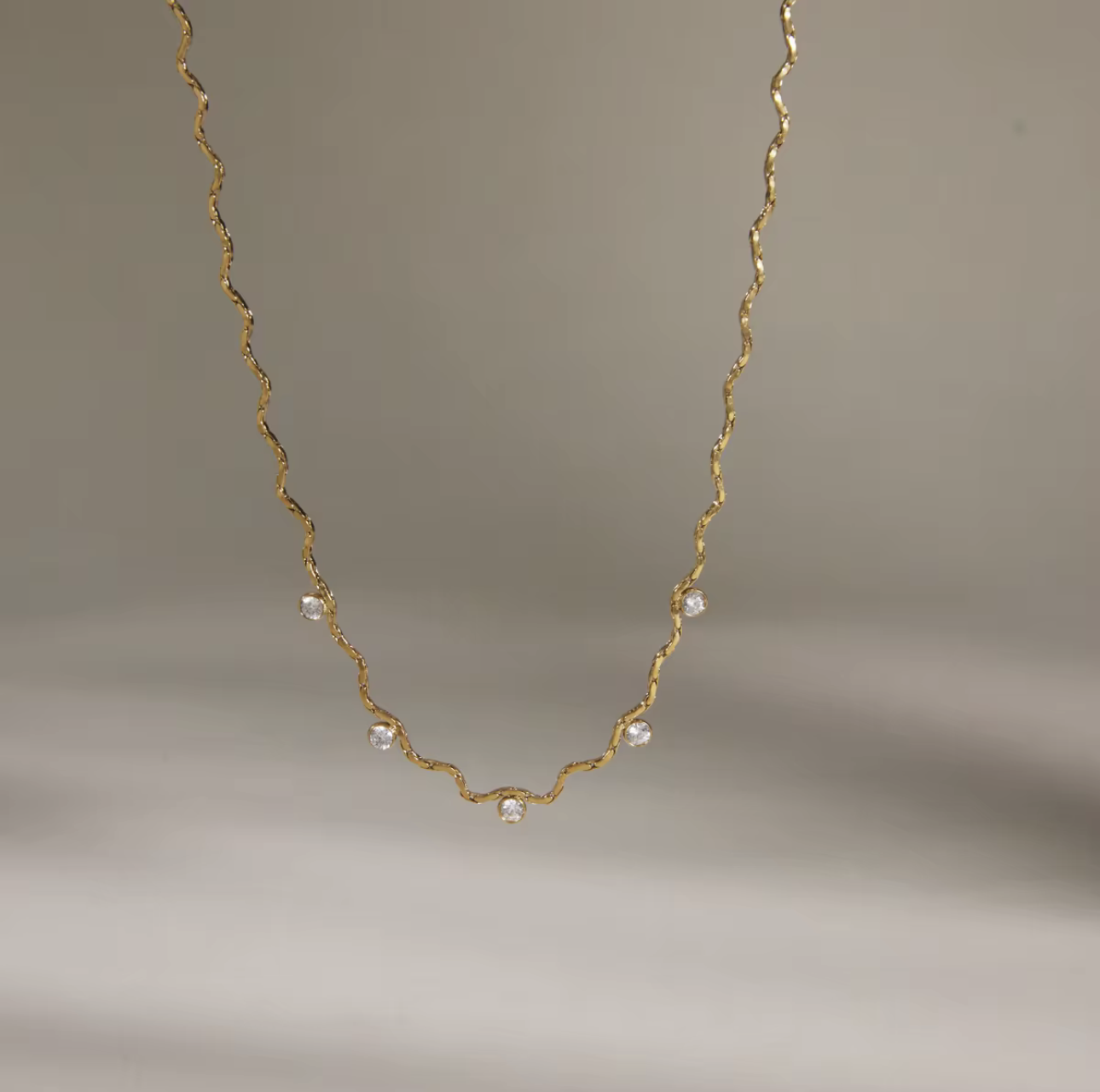 A delicate gold chain necklace with small clear faceted beads spaced along the wavy chain, photographed against a soft neutral gray background.