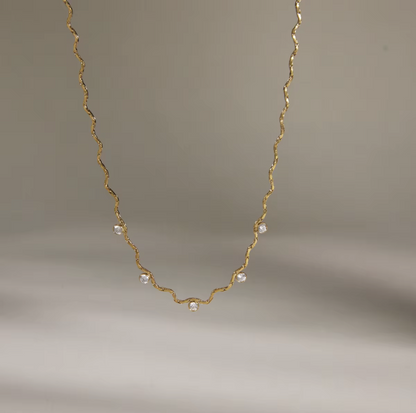 A delicate gold chain necklace with small clear faceted beads spaced along the wavy chain, photographed against a soft neutral gray background.