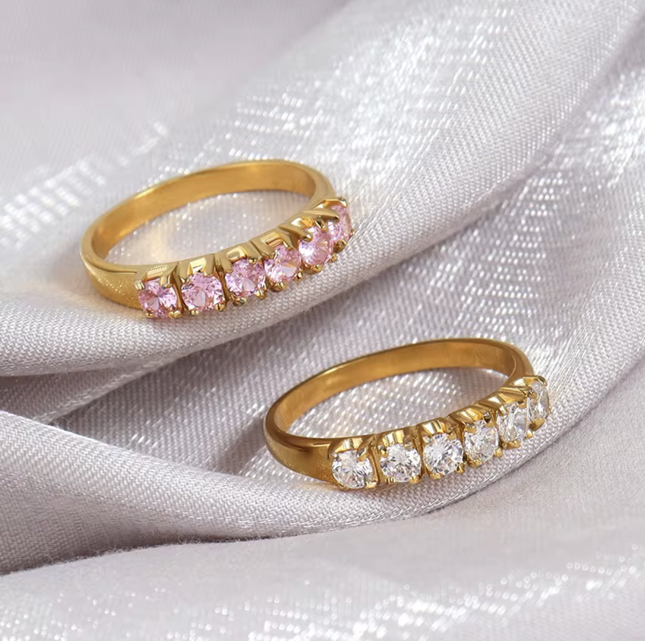 Two gold rings with pink and clear gemstones on a white fabric background